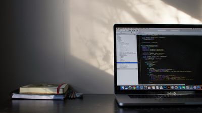 A MacBook on a desk showing code in an editor, a stack of notebooks beside it, and soft afternoon light catching the wall behind